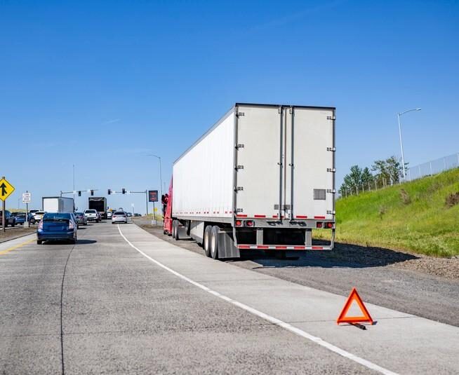 A large white semi-truck pulled over on the side of a busy freeway with a warning triangle, indicating trucking hazards.